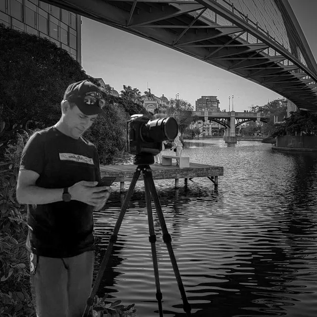 Arnold checking his phone beside a canal, camera mounted on a tripod under a footbridge