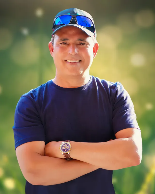 Arnold Petersen outdoors, arms folded and smiling, a cap and mirrored sunglasses pushed up on the brim