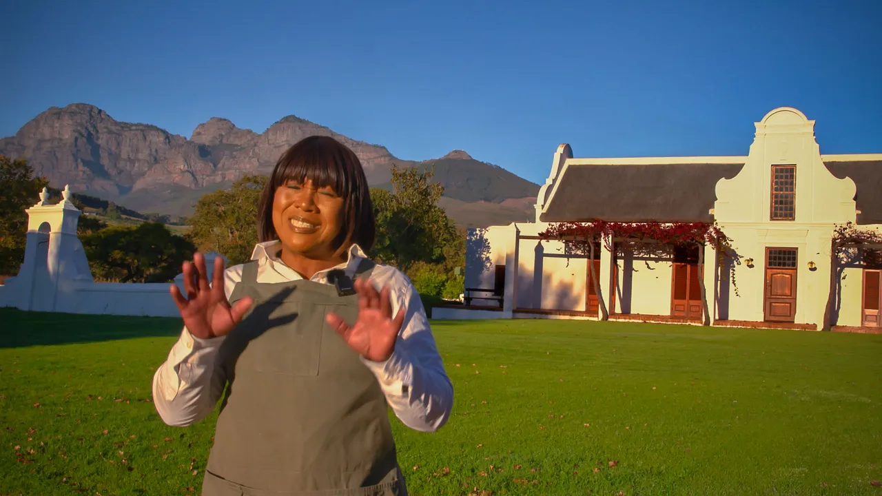 A woman in a work apron laughing with both hands raised on the lawn, a whitewashed Cape Dutch werf and the Simonsberg ridge behind her