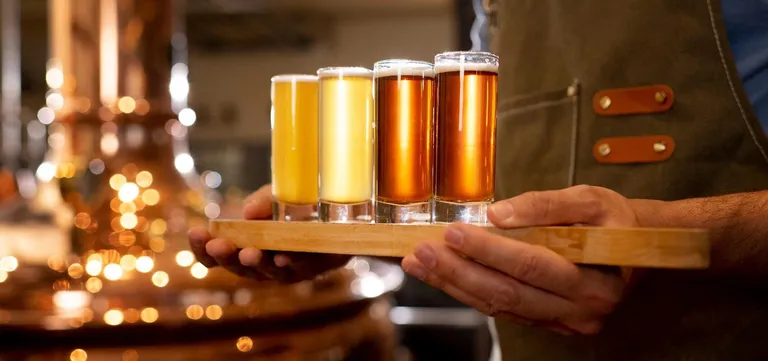 A tasting flight of four Belgian beers on a wooden paddle, carried past a copper still