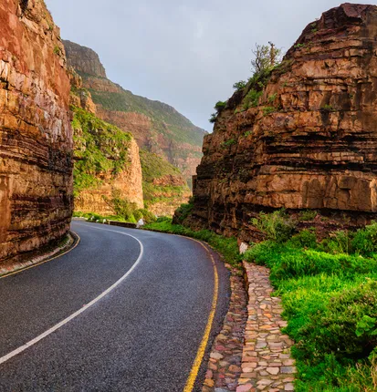 A photograph from the book: Chapman's Peak Drive curving between layered red sandstone cliffs, a cobbled kerb and green scrub along its edge