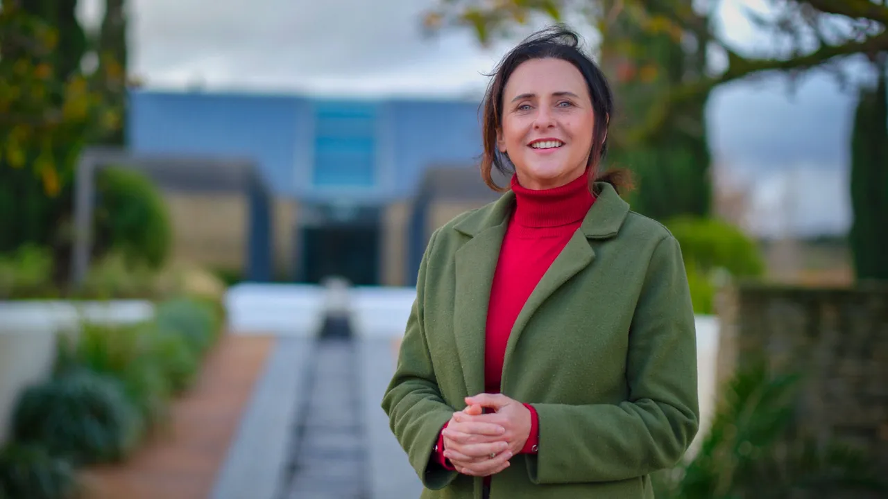 A woman in a green coat and red polo neck standing in the winter garden, the low glass pavilion and its water channel stretching away behind