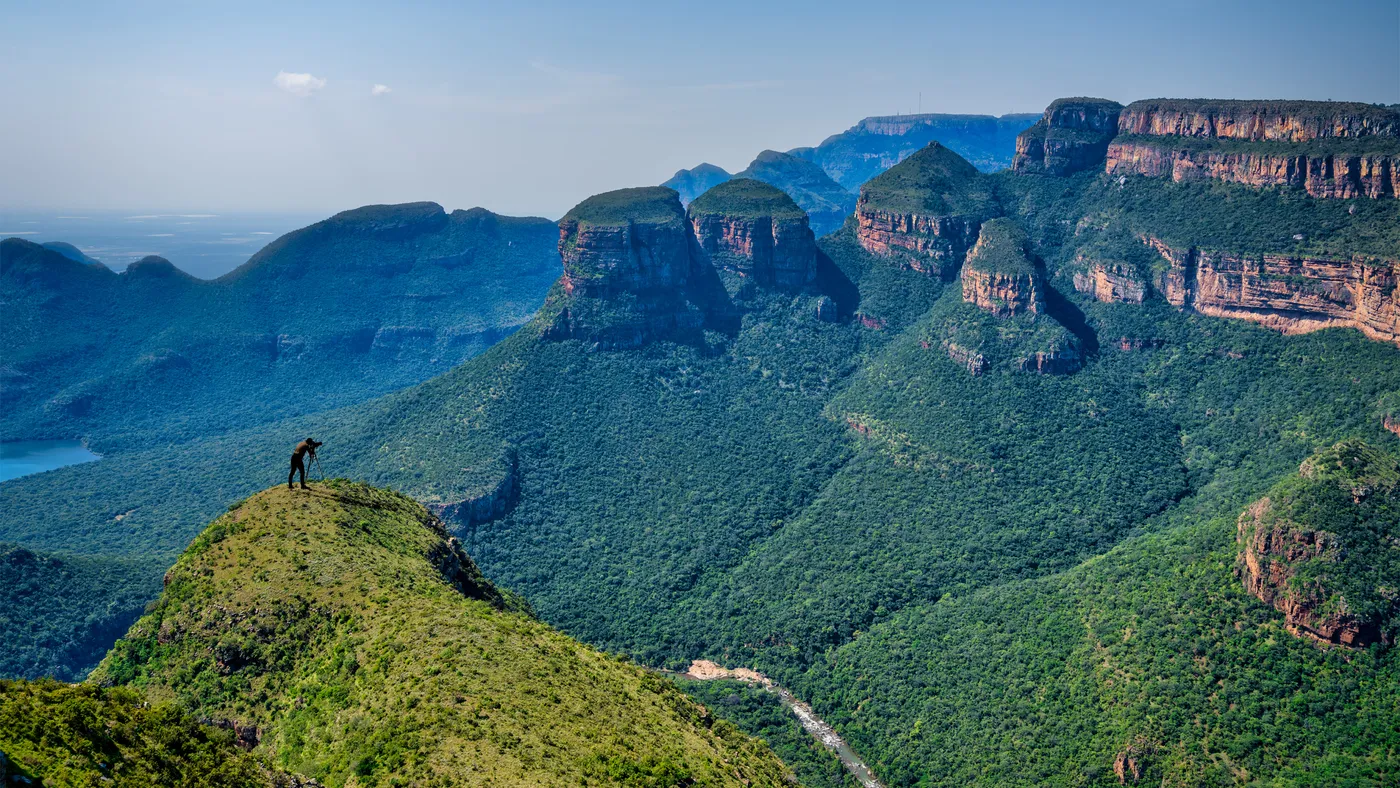 A lone photographer at a tripod on a grassy outcrop high above the Blyde River Canyon, the Three Rondavels across the gorge