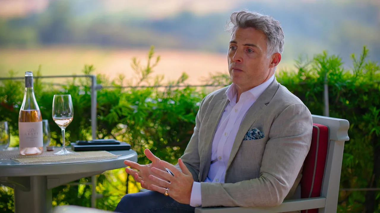 A man in a pale linen jacket gesturing as he speaks, a bottle of rosé and two glasses on the table beside him, the Helshoogte slopes soft behind
