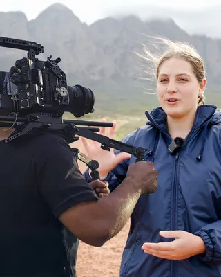A camera operator with a shoulder-mounted cinema rig filming a young woman in a navy rain jacket, wind in her hair, a clip-on mic on her collar, speaking with both hands raised before a misty mountain range.