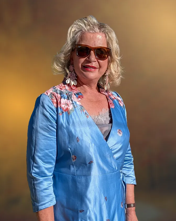 A blonde woman in dark sunglasses and long drop earrings, wearing a blue wrap dress with a floral shoulder, against a warm gold backdrop