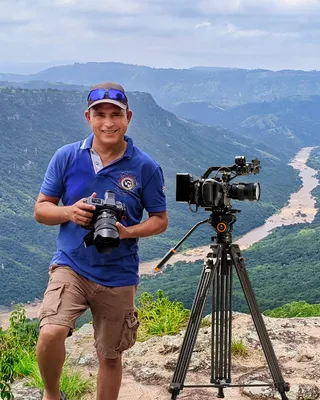 A photographer in a blue polo and cap holding a stills camera beside a cinema camera on a tripod, on a clifftop above a deep green river gorge winding into the distance