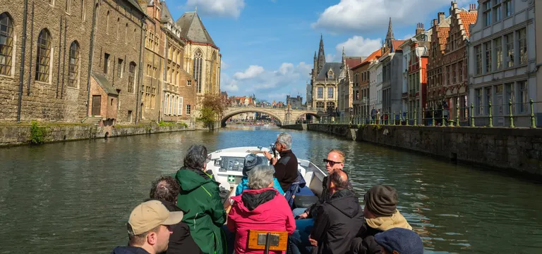 Passengers aboard an open canal boat on the Leie in Ghent, approaching a stone bridge lined with historic houses