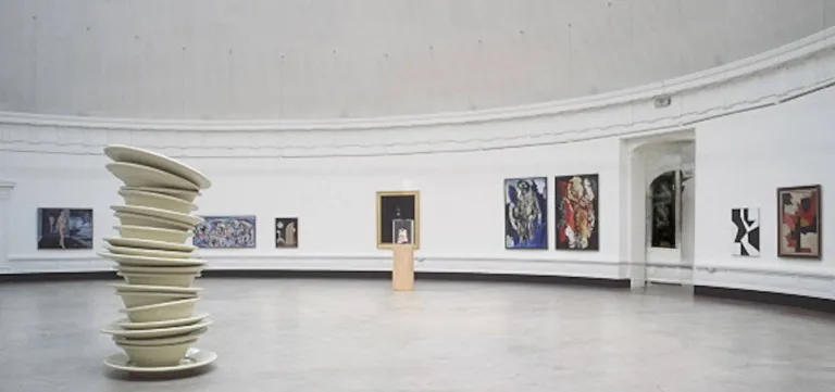 A curved white gallery hung with modern paintings, a tall stack of ceramic bowls standing as a sculpture at its centre