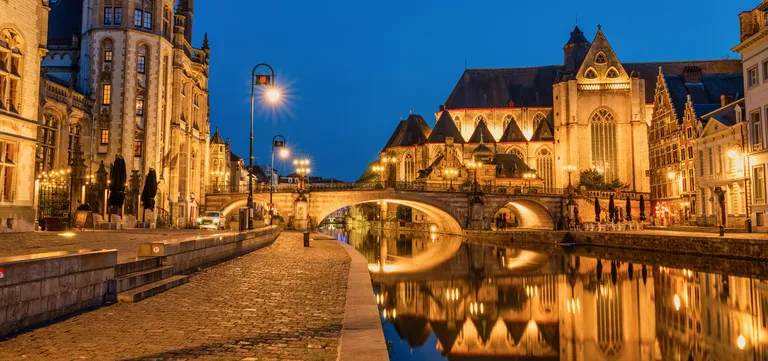 The floodlit arch of Saint Michael's Bridge and Saint Michael's Church reflected in the Leie at night, seen from Ghent's cobbled quayside