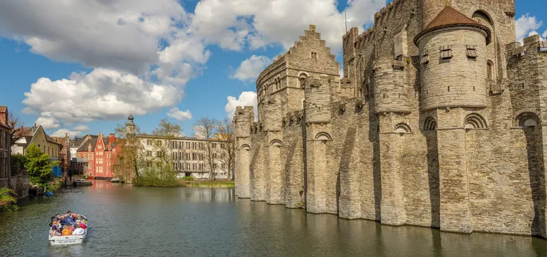 The moated stone walls and turrets of Gravensteen Castle in Ghent, with a small tour boat passing beneath them