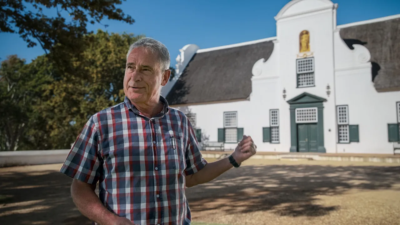 A winemaker in a red checked shirt talking with his hands on the lawn outside the white gabled Groot Constantia manor house