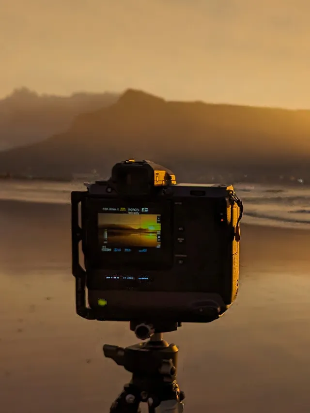 A caged mirrorless camera counts down a long exposure on wet sand, headland silhouetted in sunset haze