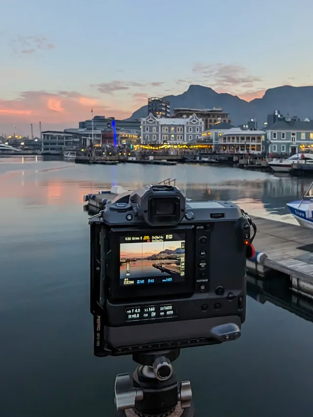 A tripod-mounted camera faces the harbour at dusk, Table Mountain behind the lit Victorian buildings