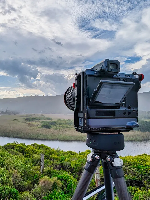 A Fujifilm GFX on a carbon tripod turned toward a river winding through reed beds under heavy cloud