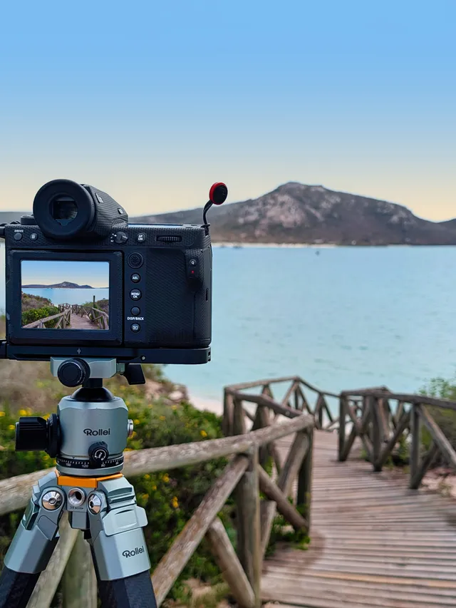 A camera on a Rollei tripod beside a wooden boardwalk descending to a turquoise lagoon and hill
