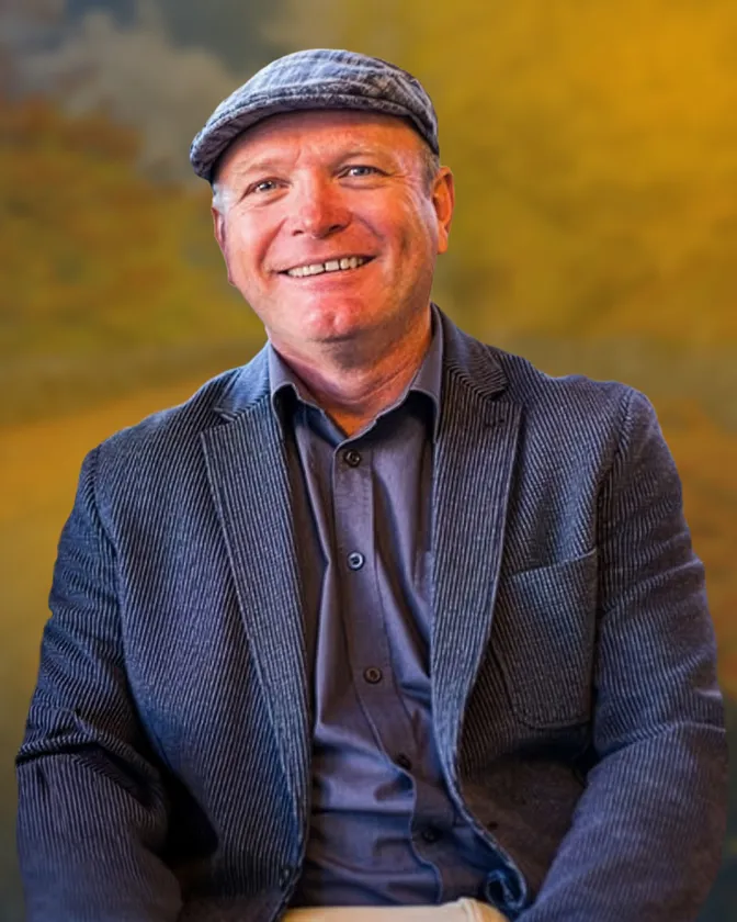 A smiling man in a grey flat cap and blue-grey corduroy jacket over a dark shirt, against a warm autumn-toned backdrop