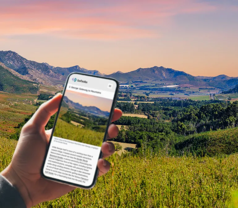 Hand holding a phone open to the GeoParallax Garden Route guide, with the Outeniqua mountains near George behind it