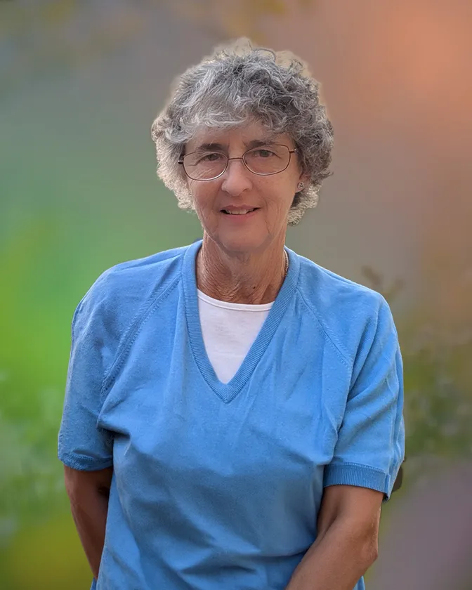 A woman with short grey curls and wire-rimmed glasses in a blue V-neck top, against a softly blurred garden background