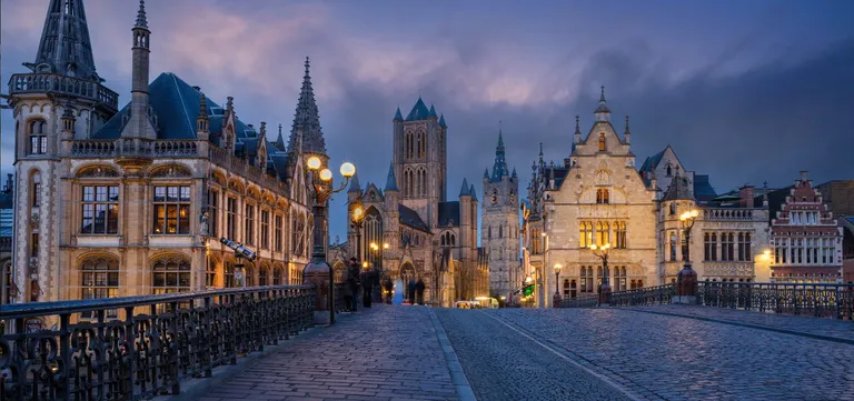 Saint Nicholas' Church and the Belfry beyond it at dusk, seen from the cobbled deck of Saint Michael's Bridge in Ghent